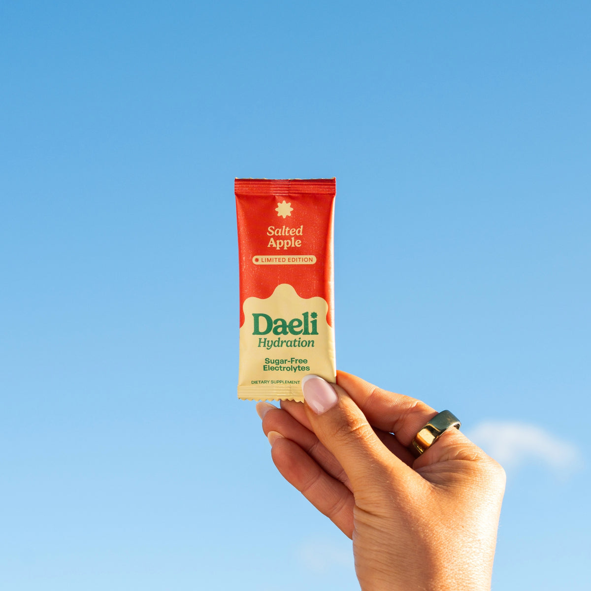 Hand holding Daeli Salted Apple electrolyte sachet against a bright blue sky - refreshing hydration for everyday energy.