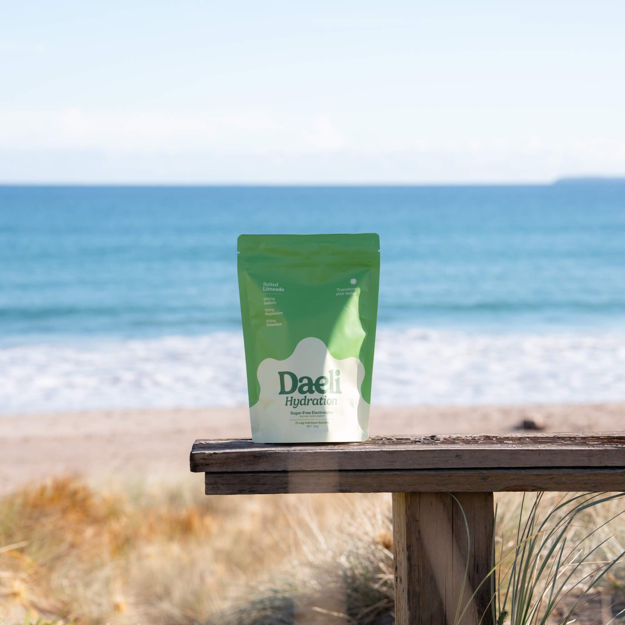 Daeli Hydration Salted Limeade electrolyte pouch standing on a beachside bench with ocean in the background.