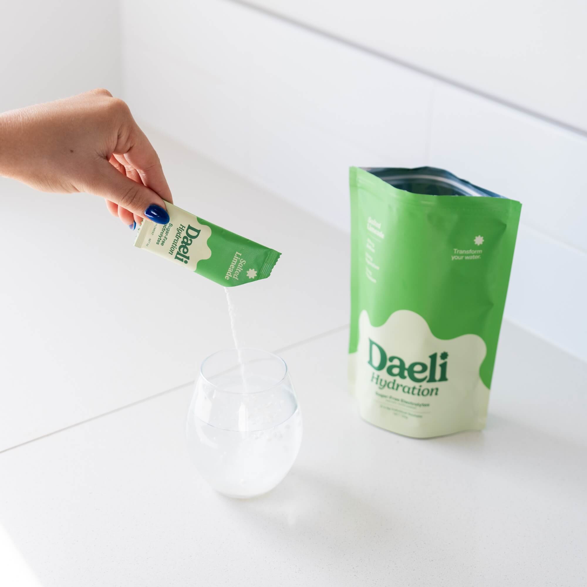Hand pouring Daeli Hydration Salted Limeade electrolyte powder into a glass of water on a white kitchen counter - hydration made easy.