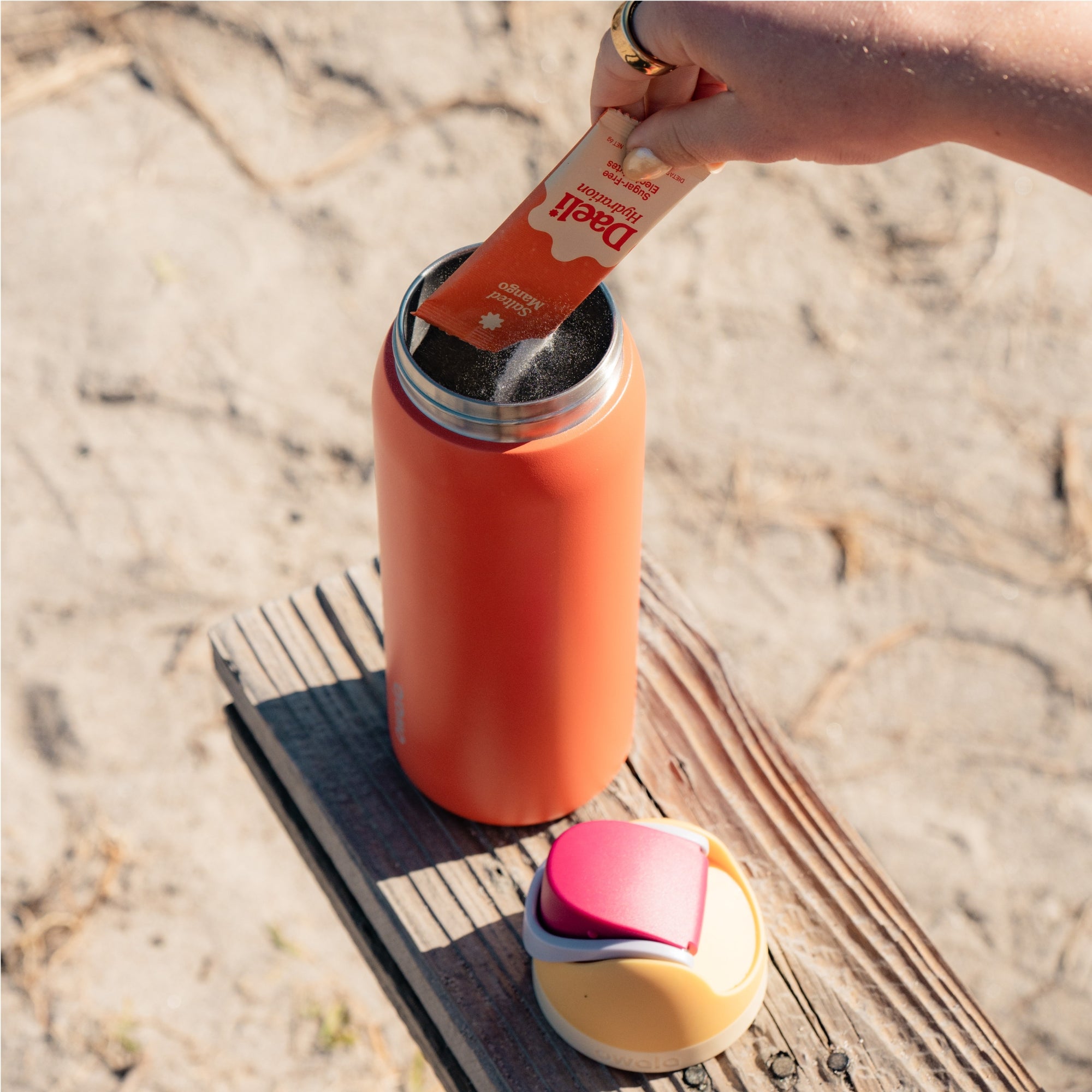 Hand pouring Daeli Hydration Salted Mango electrolyte powder into an Owala FreeSip drink bottle sitting on a bench.
