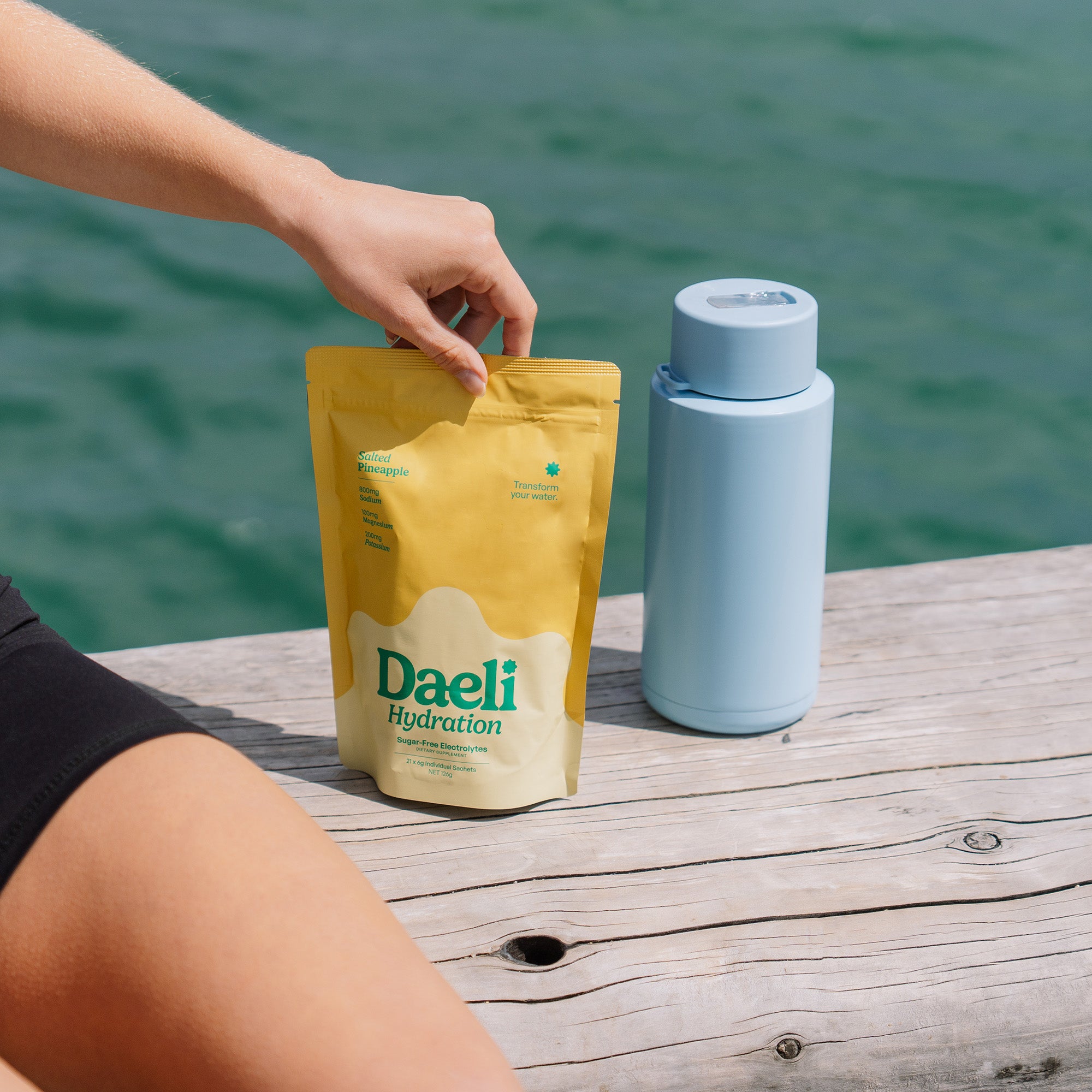 Person holding Daeli Hydration Salted Pineapple electrolyte pouch on a dock beside a blue water bottle - refreshing hydration for life on the move.
