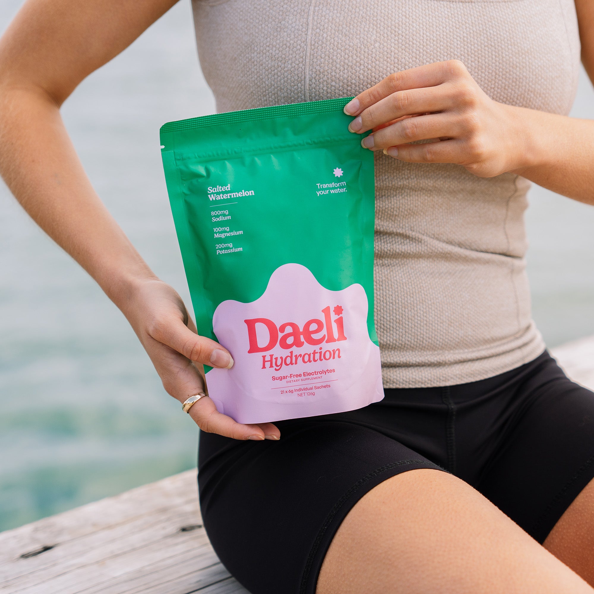 Person wearing activewear holding Daeli Salted Watermelon electrolyte pouch while sitting by the water - clean hydration for an active lifestyle.