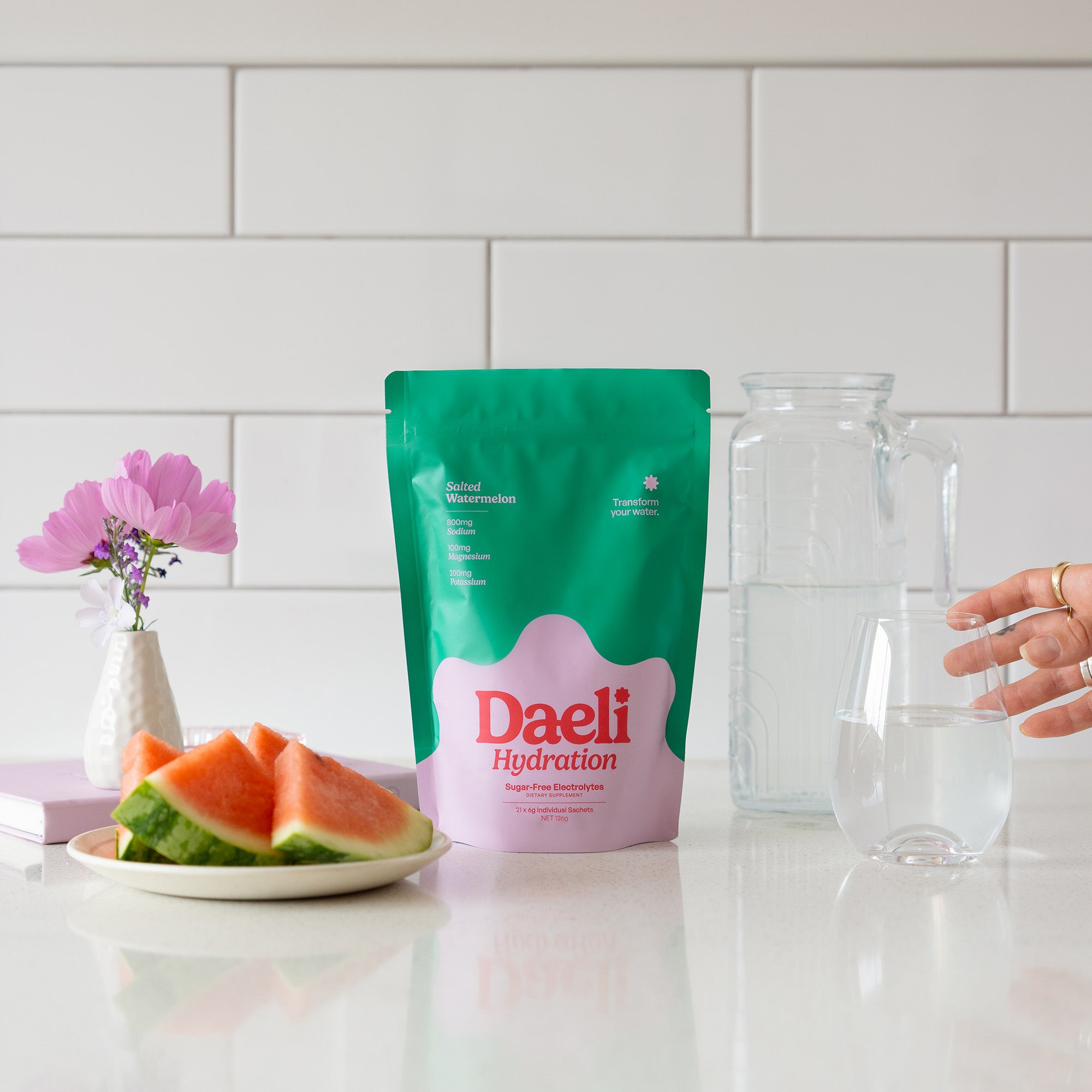 Daeli Hydration Salted Watermelon electrolyte pouch on a kitchen bench with sliced watermelon, a glass of water and fresh flowers - everyday hydration made simple and beautiful.