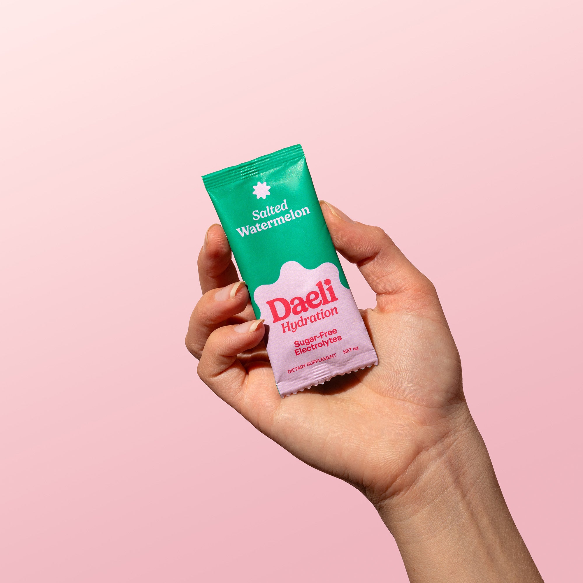 Hand holding Daeli Hydration Salted Watermelon electrolyte sachet against a soft pink background - on the go sugar-free hydration.