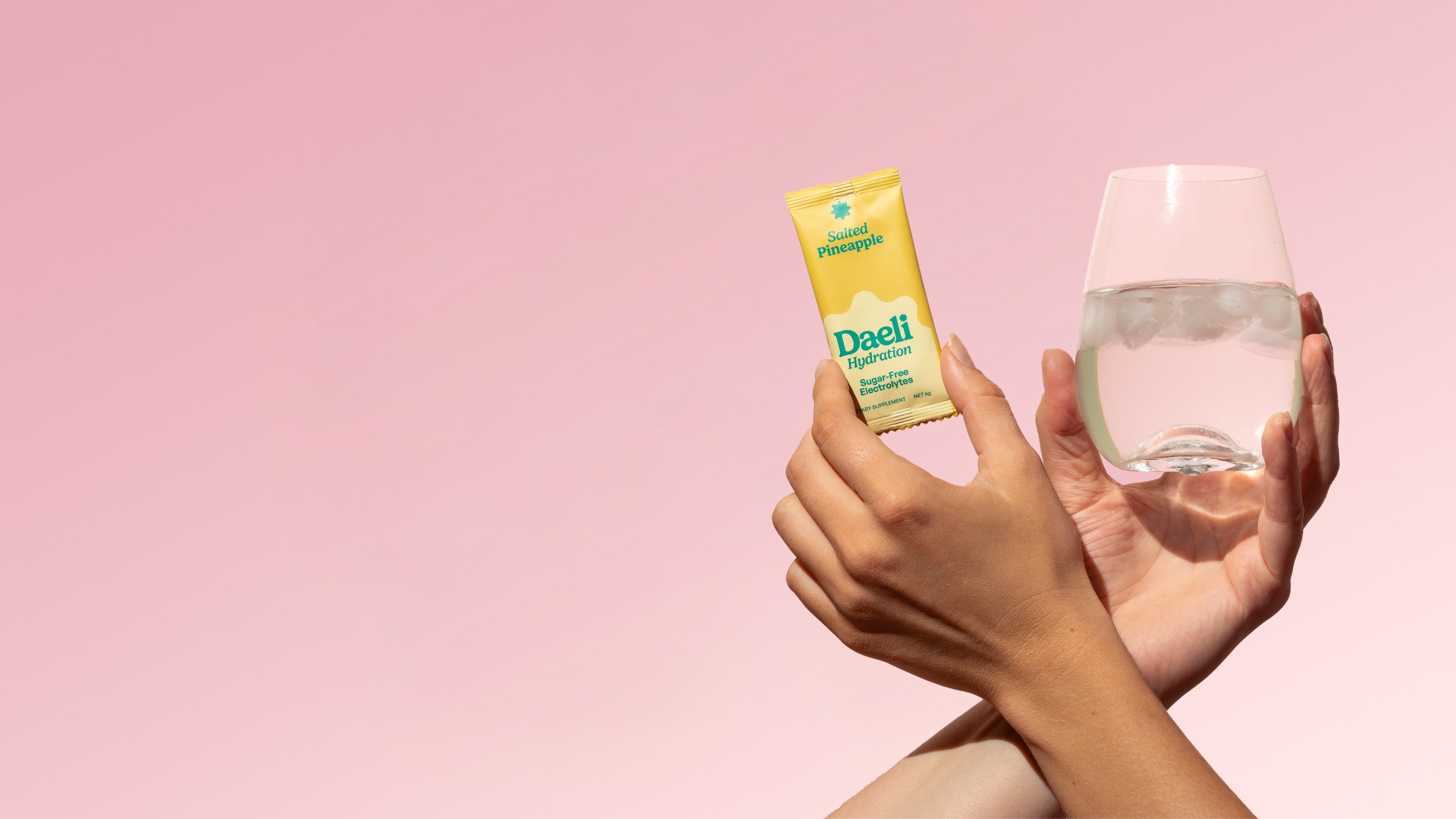 Friends of Daeli - Hand holding Daeli Hydration Salted Pineapple electrolyte sachet and glass of iced water mixed with Daeli Hydration against a soft pink background - refreshing daily hydration ritual, styled for desktop.