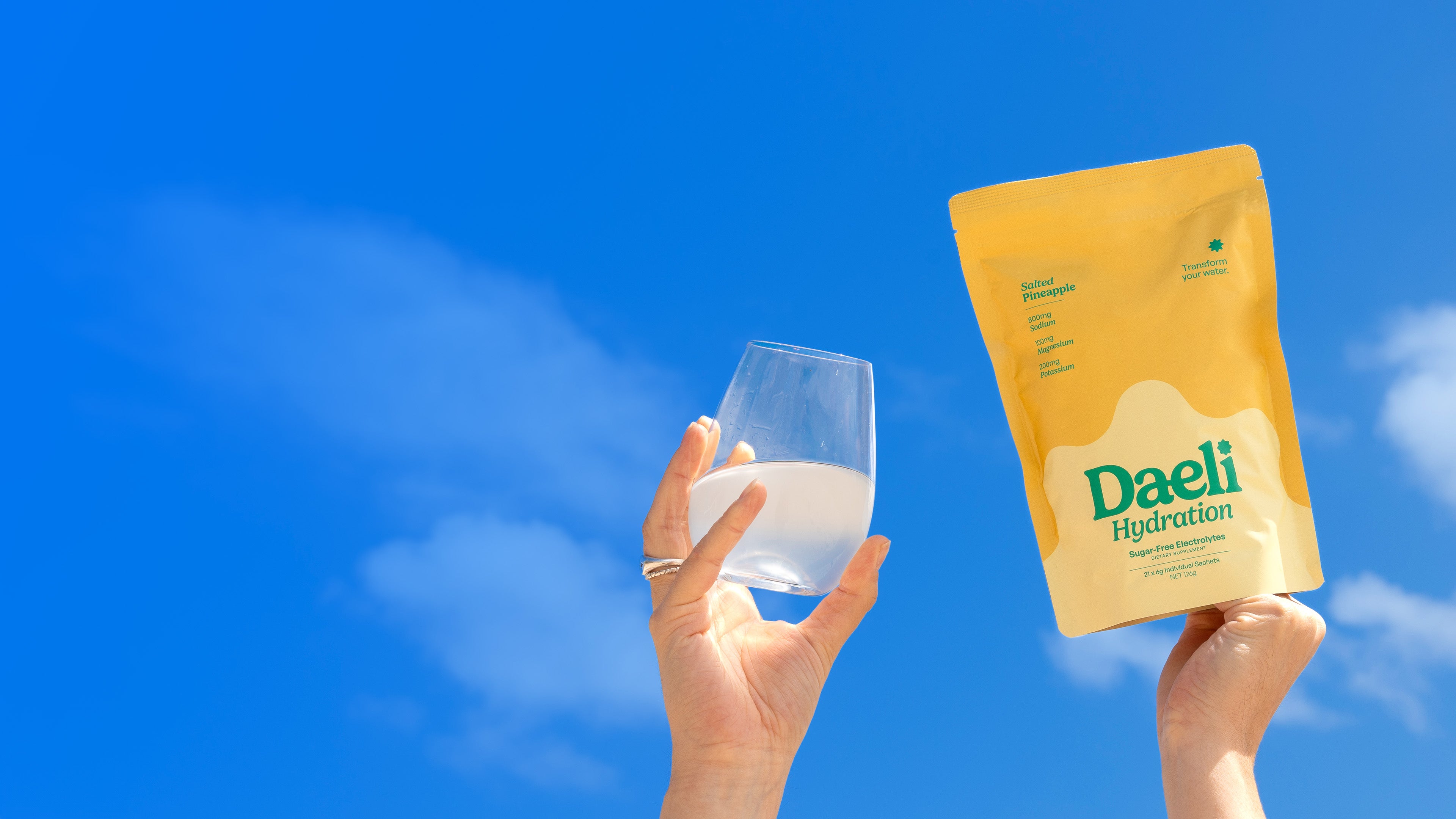 Our Range - Hand holding Daeli Hydration Salted Pineapple electrolyte powder pouch and a glass of mixed drink against a clear blue sky - sugar-free, no-nasties, travel-friendly wellness supplement, styled for desktop.