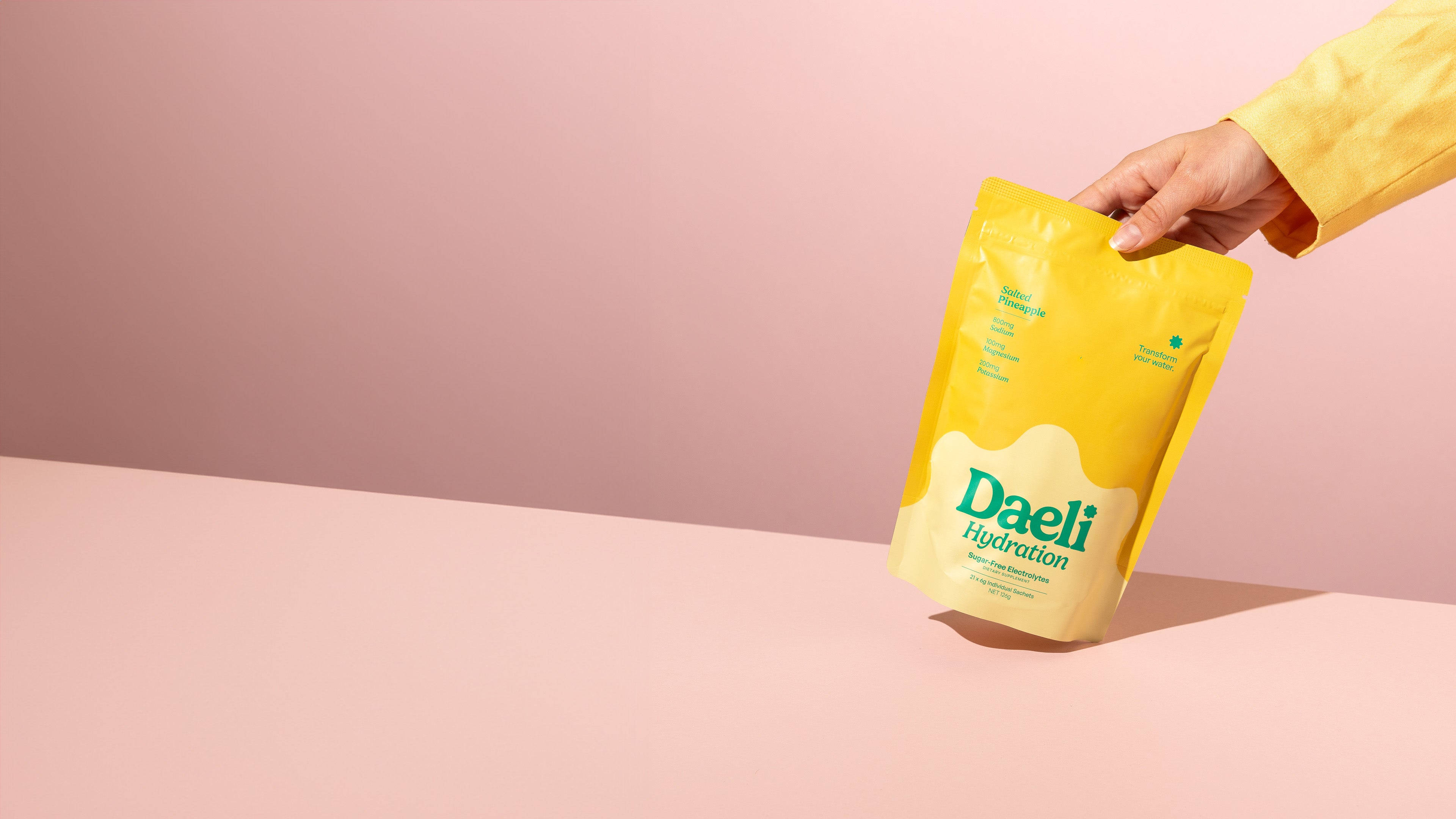 Our Story - Hand placing Daeli Salted Pineapple electrolyte pouch on a soft pink surface - clean, vibrant wellness with a daily dose of hydration, styled for desktop view.