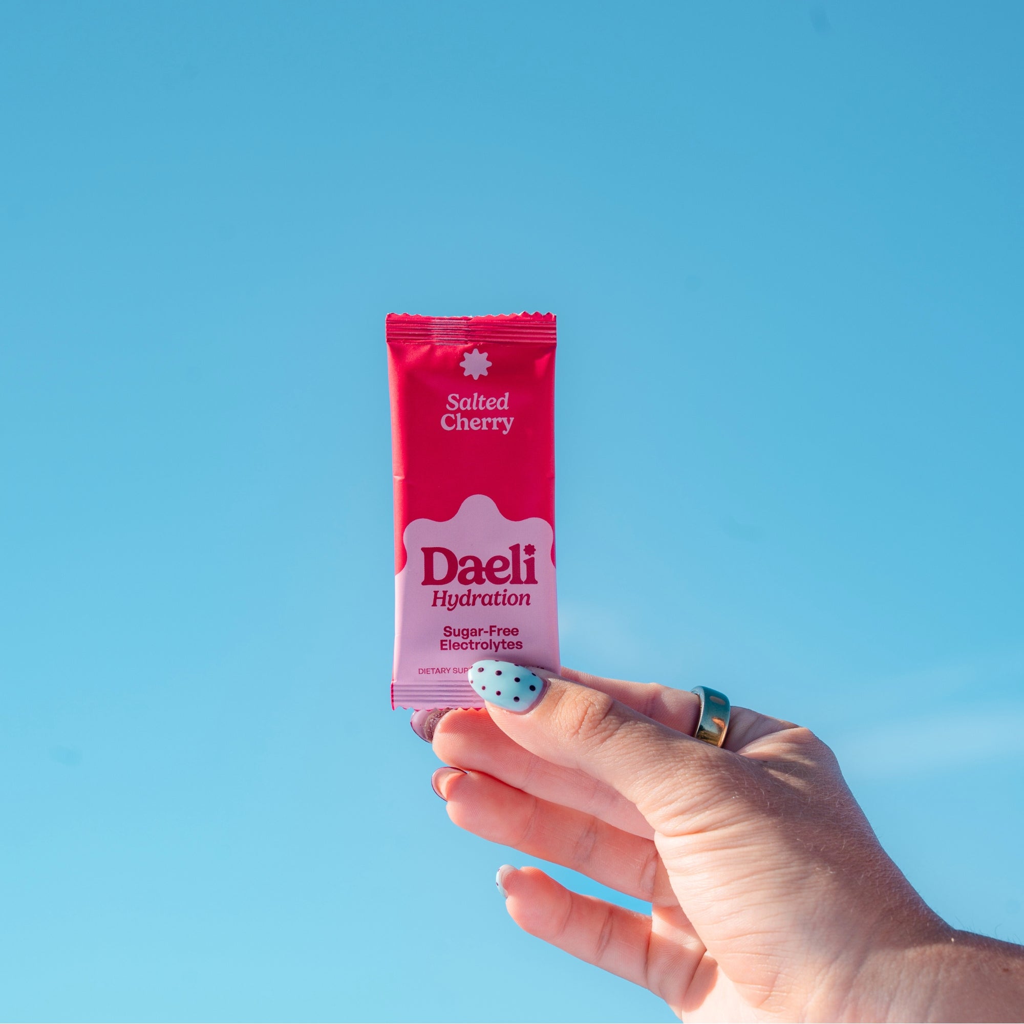 Hand holding Daeli Salted Cherry electrolyte sachet against a bright blue sky - refreshing hydration for everyday energy.